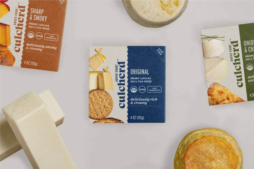 Top view of three Culcherd cheese boxes with two rounds of cheese and two sticks of butter surrounding the boxes. The far left box is an orange color of the Sharp & Smoky semi-firm flavor. The middle box is a dark blue color for the Original semi-firm cheese. The far right box is an olive green color for the Onion & Chive fresh cheese.