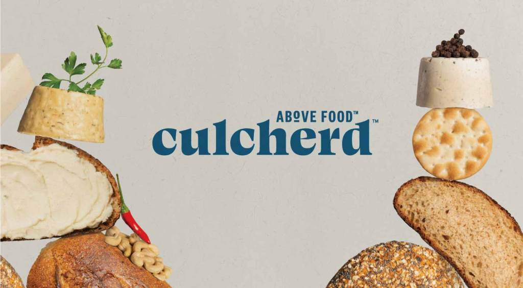 Image of the blue Culcherd logo in between two stacks of food. The left stack shows a loaf of bread on the bottom, a slice of cheese with butter spread on it on top, and a round of cheese on top of that with cilantro garnish. The right stack shows a bagel on the bottom, a slice of bread on top, a circular water cracker on top, and then a round of cheese on the very top. The cheese has a pile of black peppercorns on it.